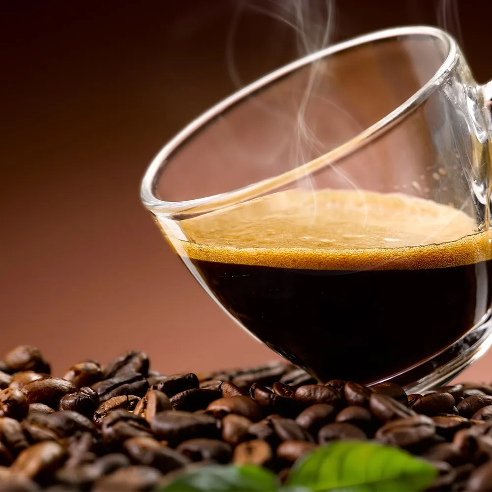 A clear glass cup of fresh espresso with a golden crema layer sitting on a pile of dark roasted coffee beans with steam rising.
