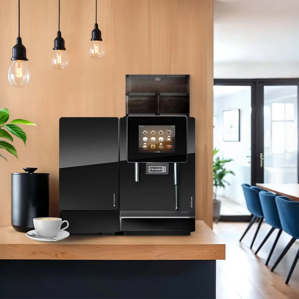 A black Franke A600 commercial espresso machine with a touchscreen and milk system on a wooden counter in a modern office kitchen with hanging lights.